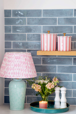 A styled interior scene with a pink patterned table lamp on a teal ceramic base, a floating wooden shelf holding two pink-and-white striped canisters, and a tray with flowers and candles against a blue-gray subway tile wall.